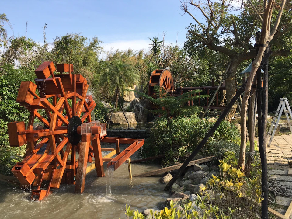 A scene featuring a wooden waterwheel over a small stream surrounded by trees, shrubs, and a clear sky.