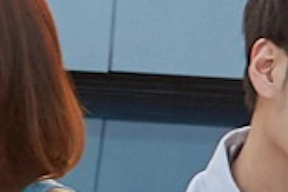 A man and a woman sit on a glass floor on a rooftop overlooking a cityscape during sunset.
