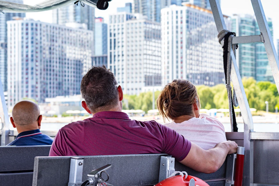 Two people sit closely on a boat, facing away, with a city skyline of tall buildings in the background.