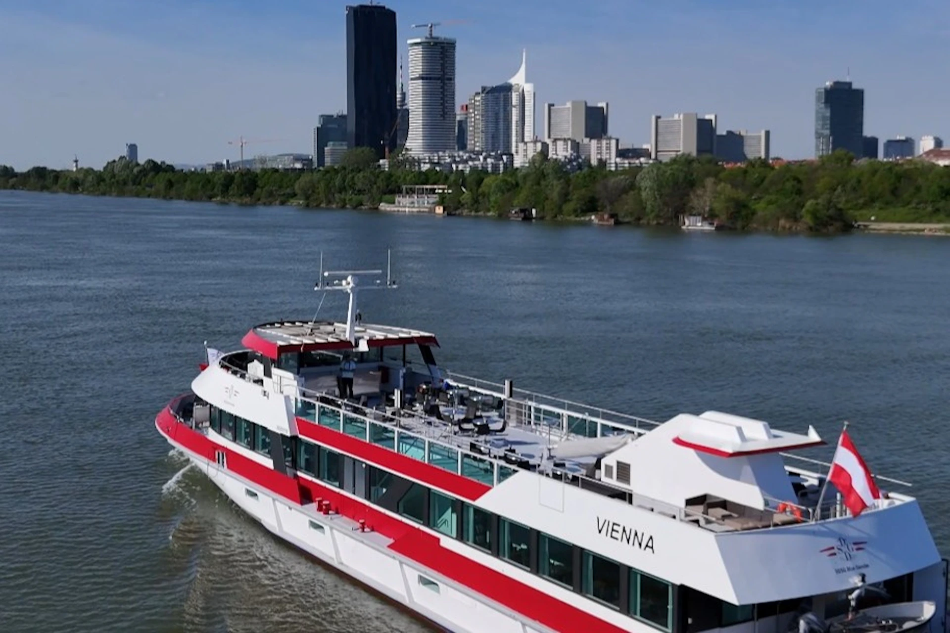 A boat named "Vienna" travels on a river with a cityscape featuring tall buildings in the background and trees lining the riverbank.