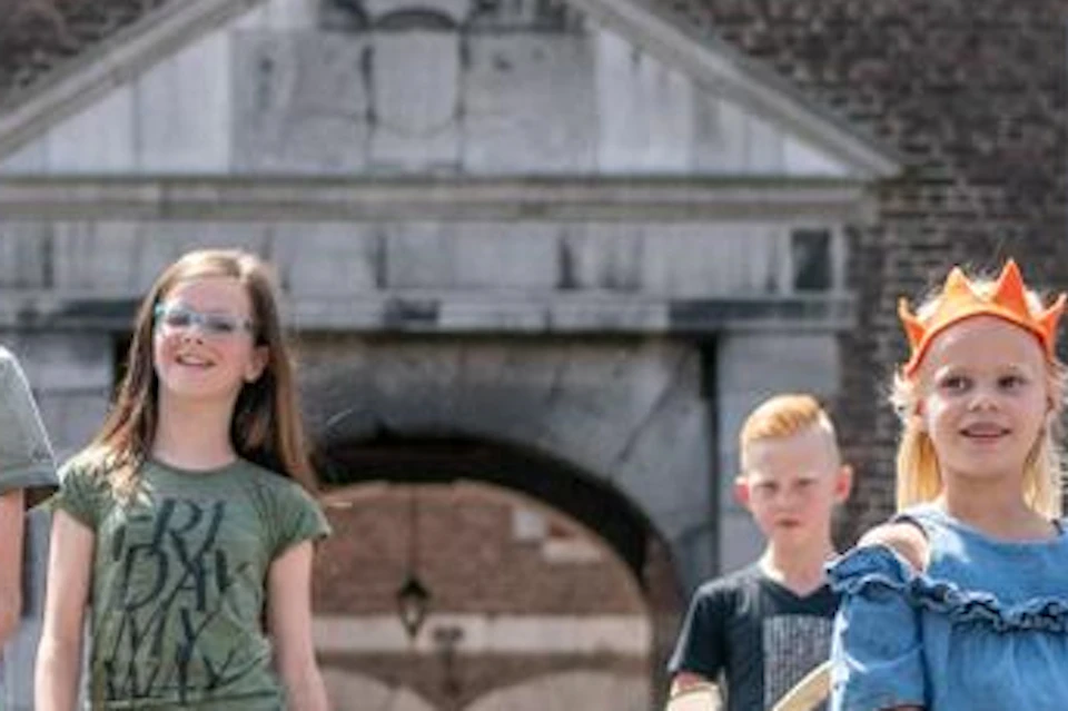 Five children wearing playful outfits and crowns walk towards the camera on a cobblestone path in front of a historic building.