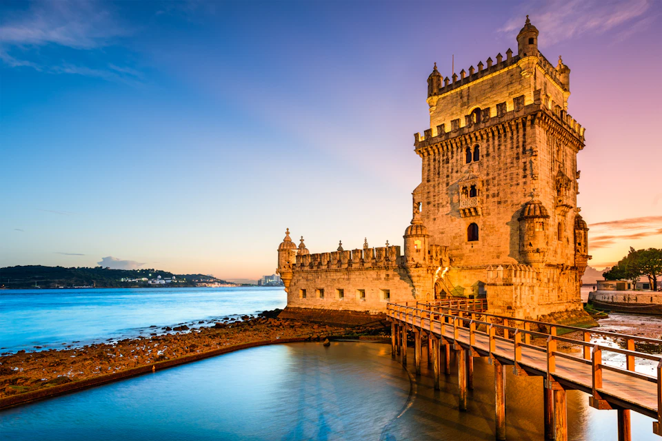 Historic stone tower with turrets by a calm waterfront at sunset, connected by a wooden walkway, under a clear sky.