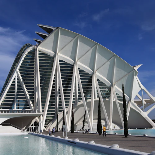 En futuristisk vit byggnad med strukturella valv, omgiven av vatten och några höga, smala träd mot en blå himmel.