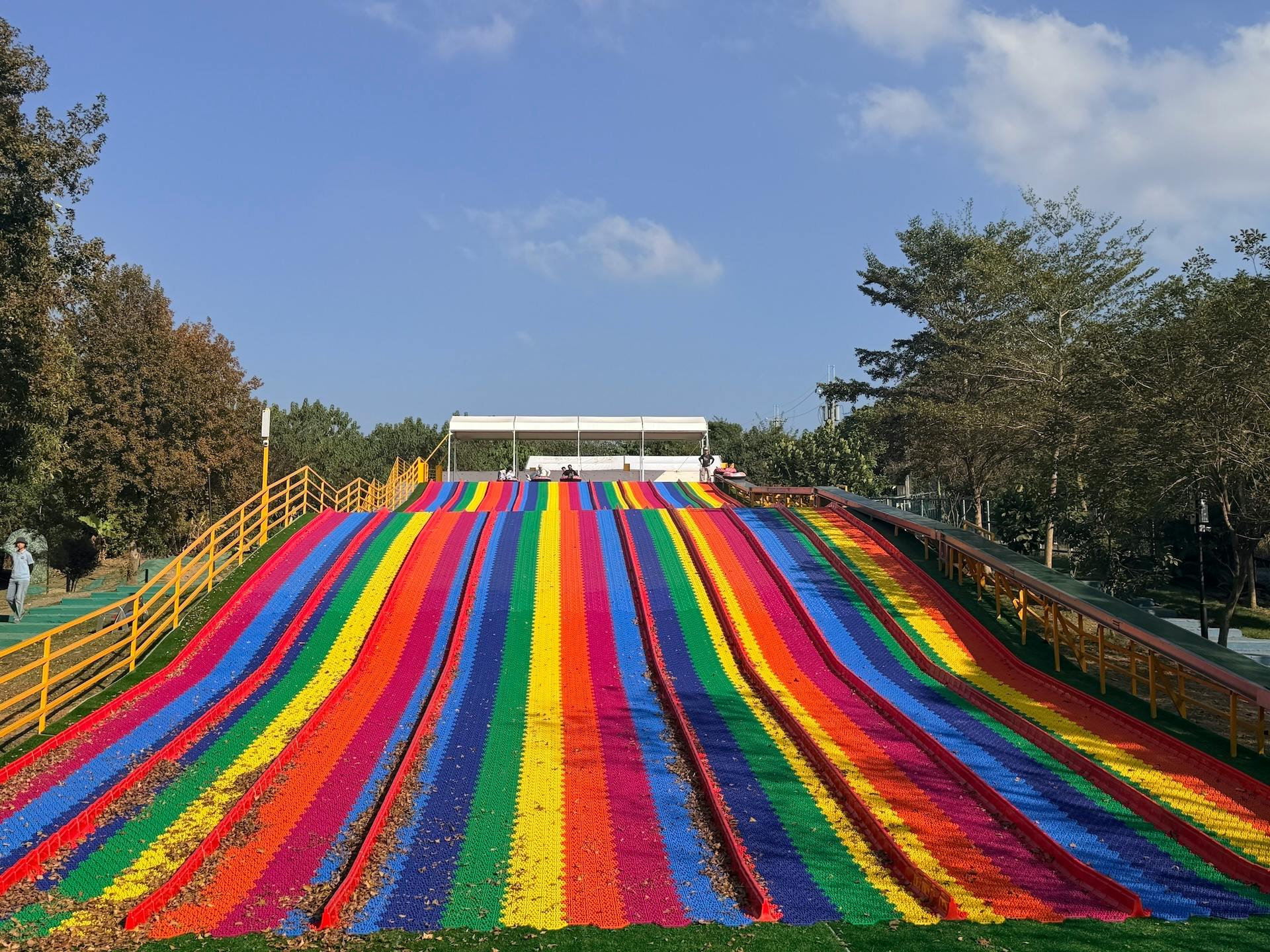 A colorful rainbow slide in a park, surrounded by trees, with a clear blue sky in the background.