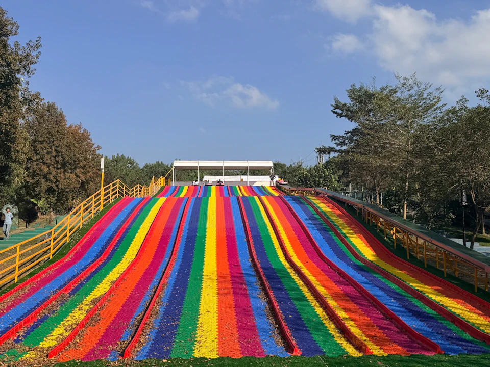 A colorful rainbow slide in a park with trees and a blue sky in the background.