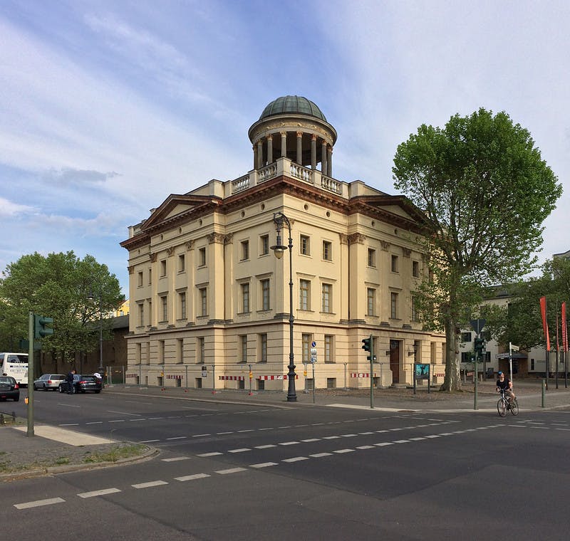 Ένα νεοκλασικό κτίριο με θολωτή ταράτσα σε μια διασταύρωση, που περιβάλλεται από δέντρα, έναν ποδηλάτη και ένα σταθμευμένο όχημα.