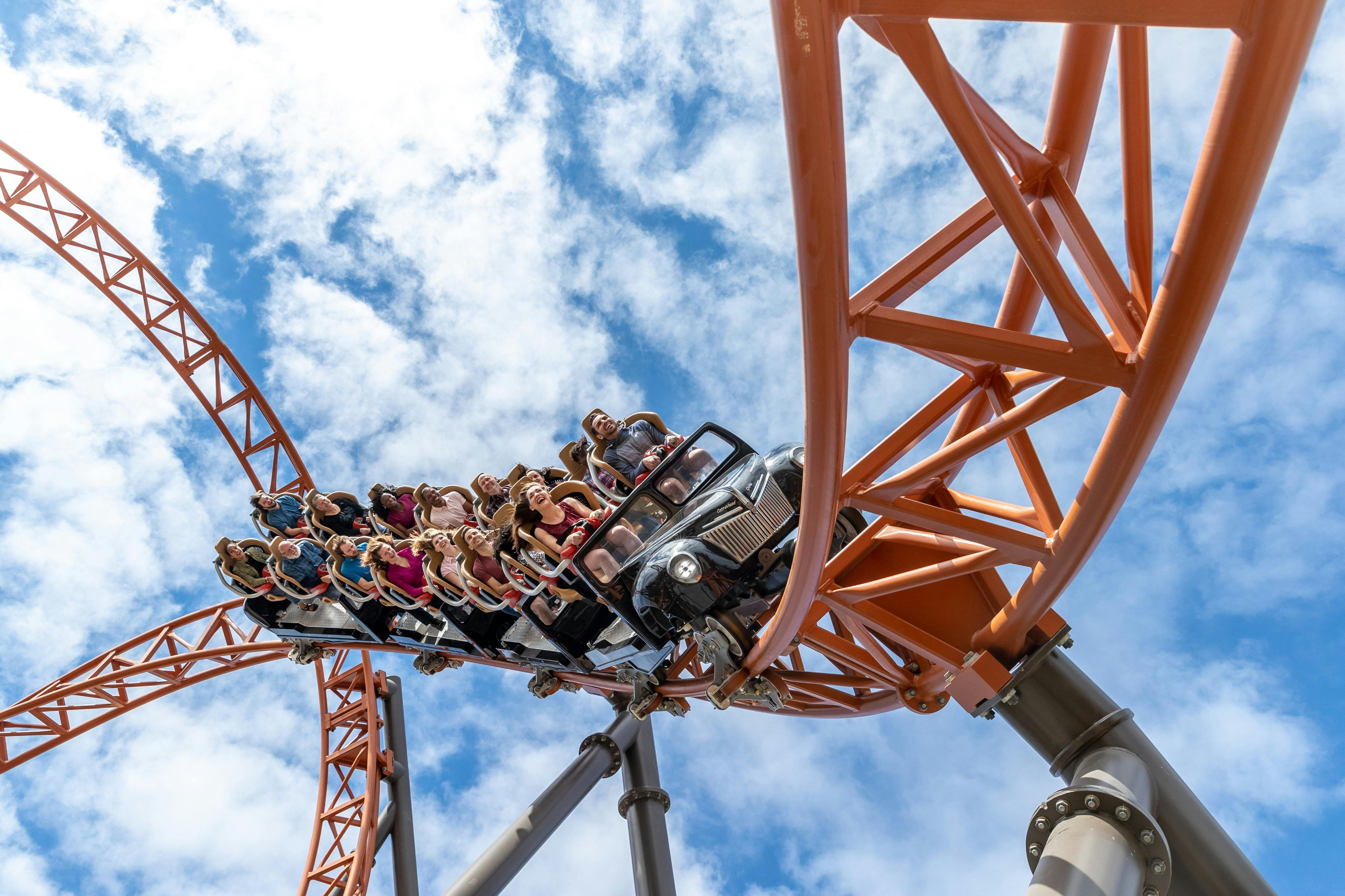 Un wagon de montagnes russes rempli de personnes sur des rails orange, capturé sur un ciel bleu avec des nuages épars.