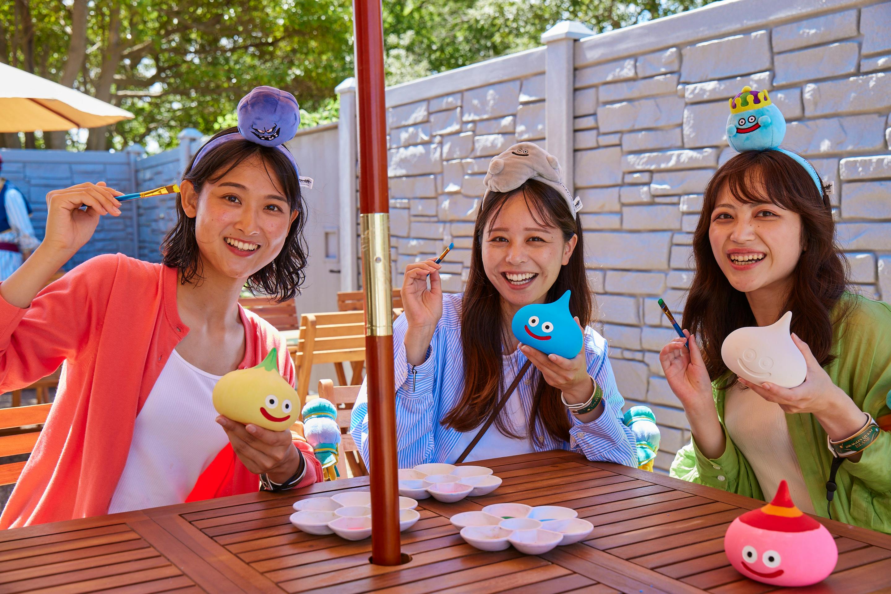 Two women sitting outdoors at a wooden table, painting small slime-shaped objects, smiling and enjoying the activity.