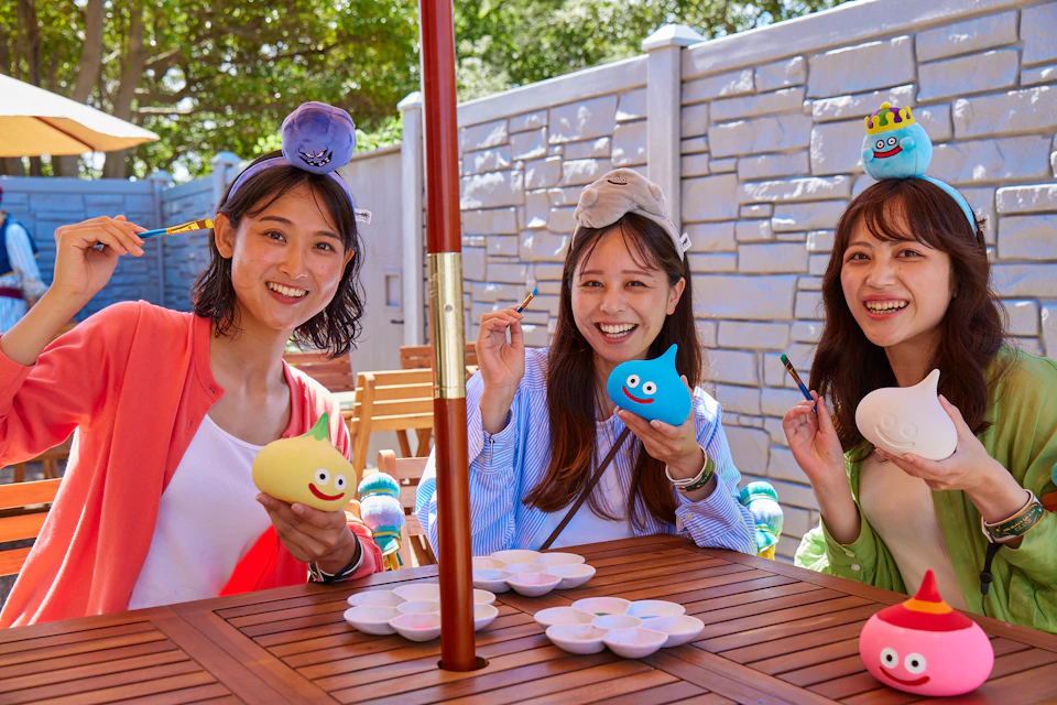 Two people painting small toys at an outdoor table under an umbrella. They smile and hold yellow and blue figures.