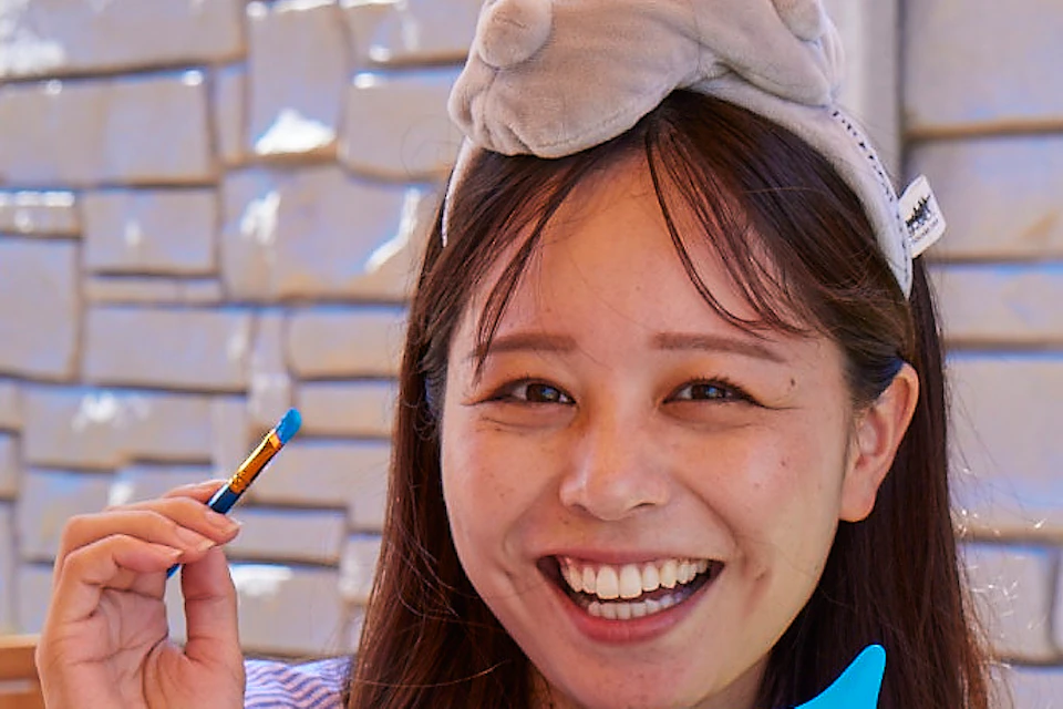 Two people painting small toys at an outdoor table under an umbrella. They smile and hold yellow and blue figures.