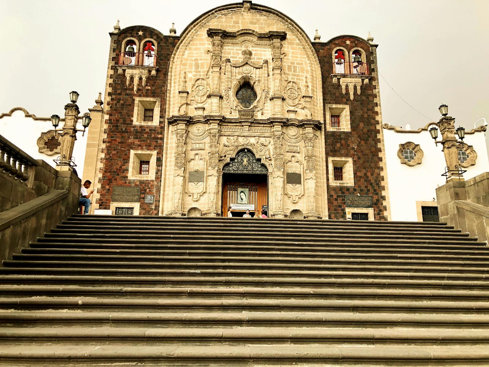 Kamenné schody vedou k ozdobné historické fasádě kostela se složitými řezbami, lemované dvěma menšími věžemi s červeně mořenými skleněnými okny.