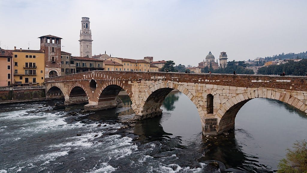 一座带有多个拱门的古老石桥横跨一条河，后面有历史建筑和一座高高的钟楼。