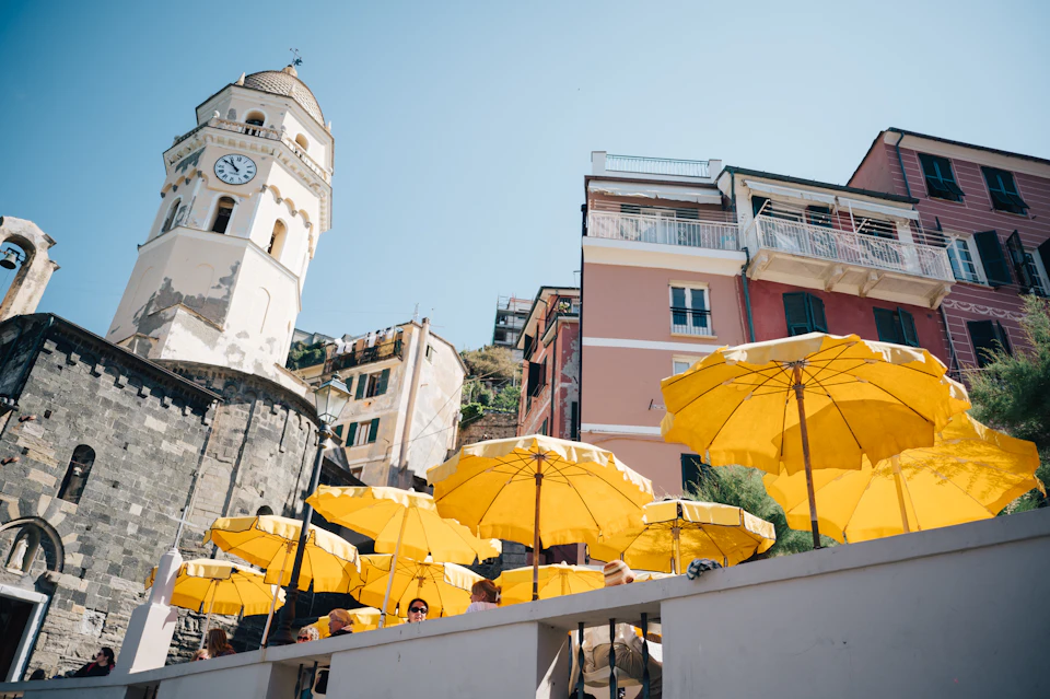 Yellow umbrellas above a terrace cafe with people, next to colorful buildings and a clock tower, under a clear blue sky.
