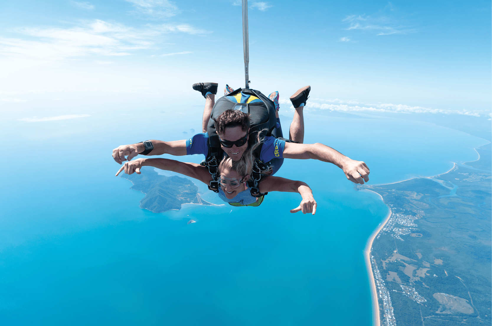 两人在清澈的蓝色海洋和沿海景观上双人跳伞，伸出双臂并佩戴安全带。