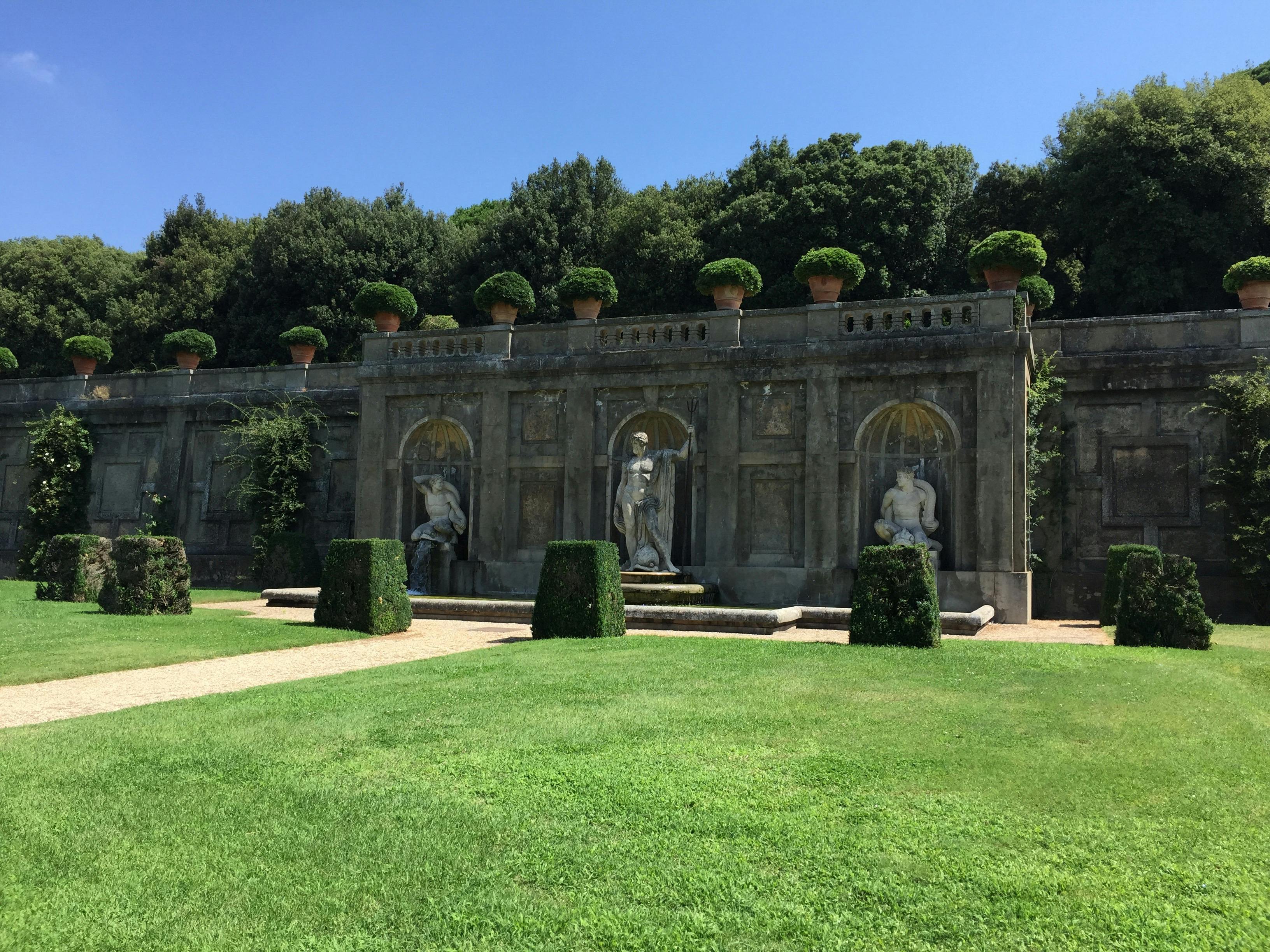 Stone statues and potted plants adorn a decorative wall in a manicured garden with hedges and a lush green lawn.