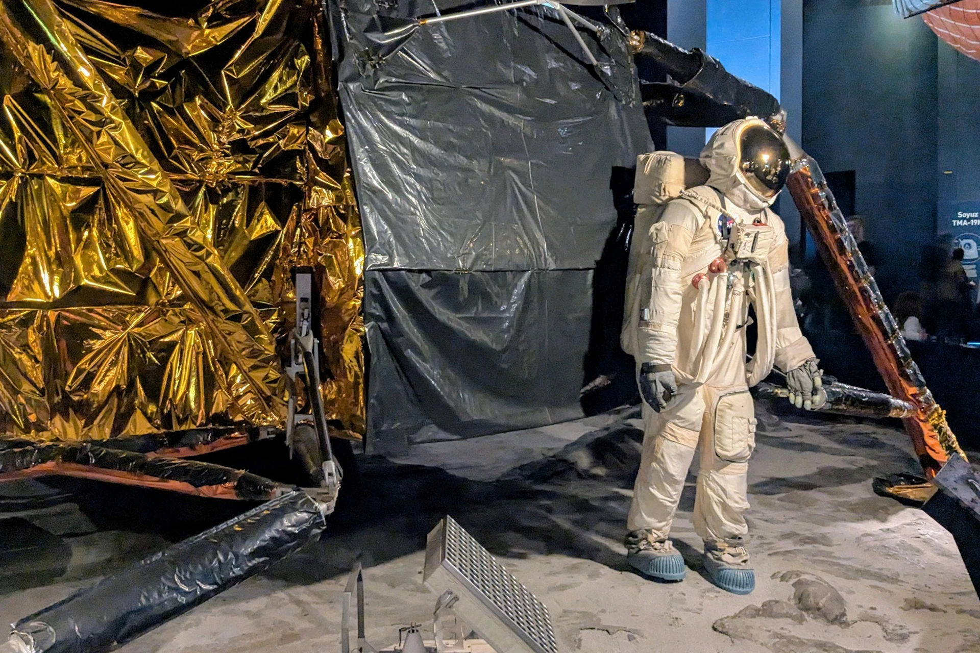 A space suit-clad mannequin stands near a replica of a lunar module, surrounded by simulated lunar surface materials.