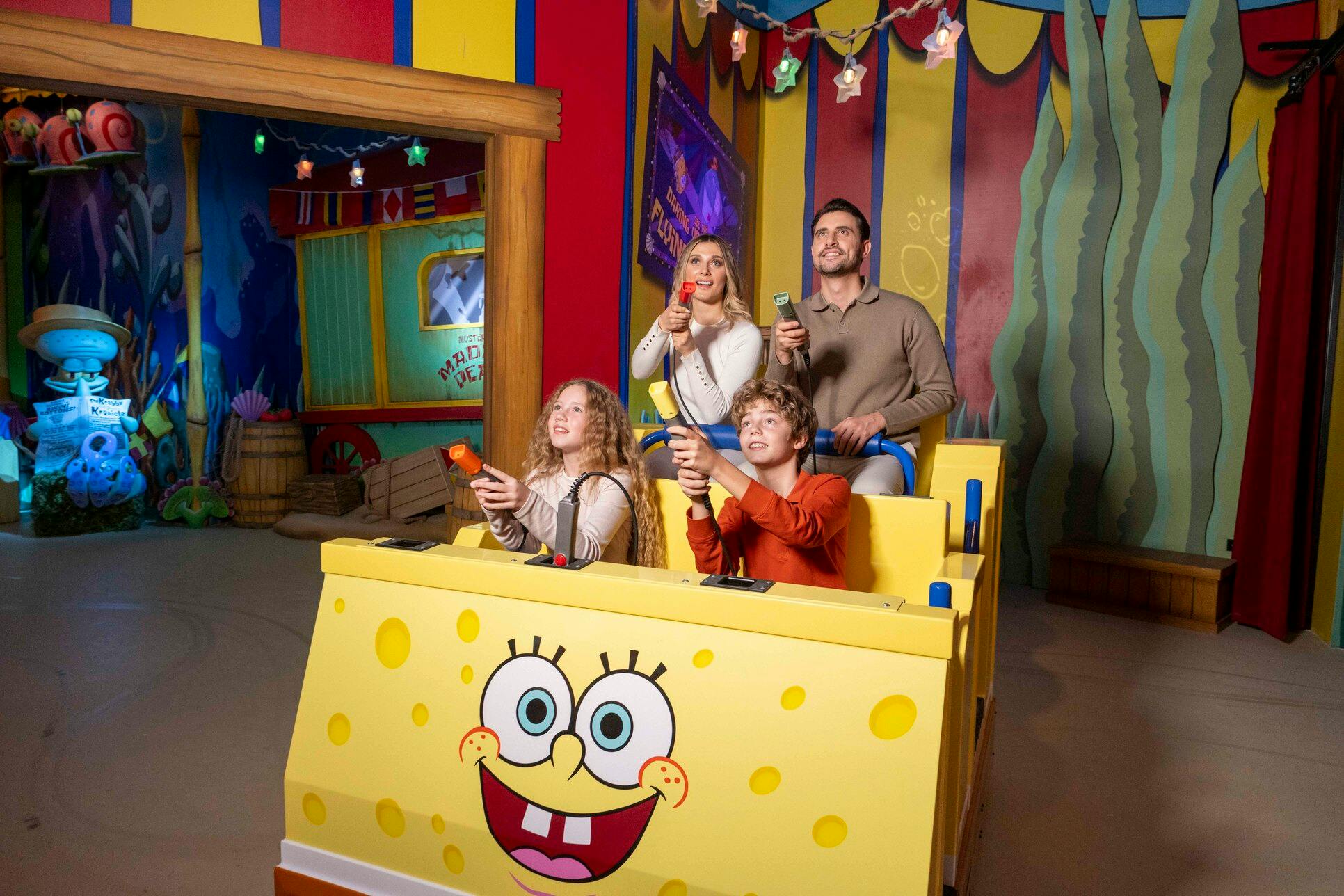 Een gezin van vier zit in een SpongeBob-attractie met lichtpistolen in een kleurrijk interieur.