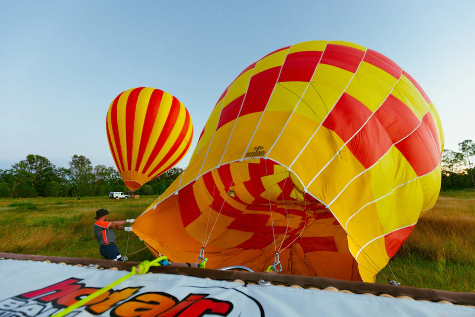 Person inflating a large yellow and red hot air balloon on the ground, with another rising in the sky behind them.