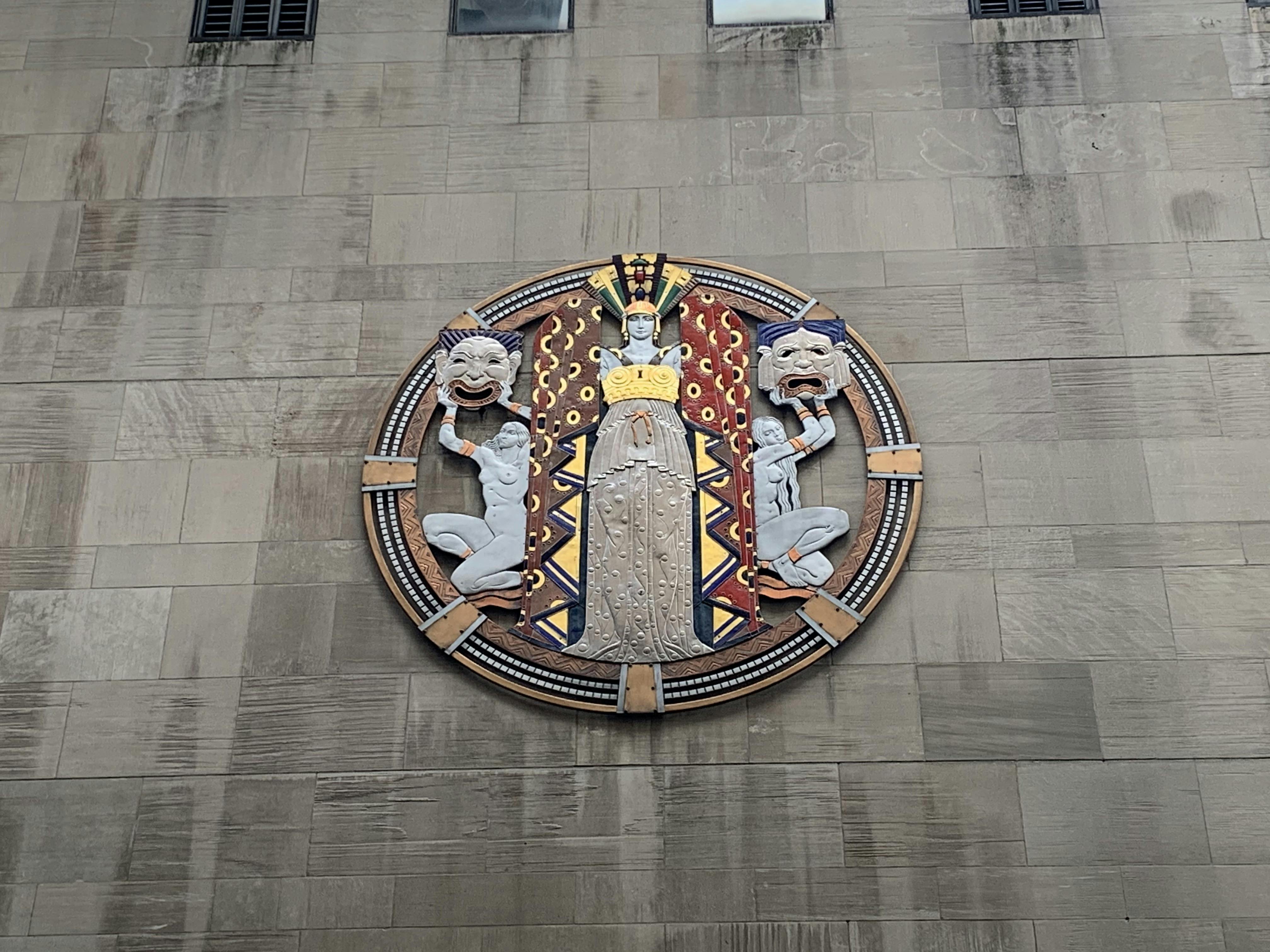 Ένα κυκλικό γλυπτό τοίχου απεικονίζει μια κεντρική φιγούρα με περίτεχνη ενδυμασία που πλαισιώνεται από δύο μικρότερες φιγούρες που κρατούν μάσκες.