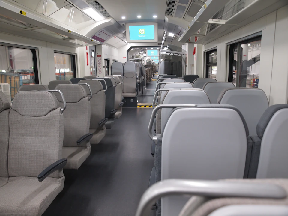 Interior of an empty train carriage with grey seats, overhead luggage racks, and screens displaying a logo and blue background.
