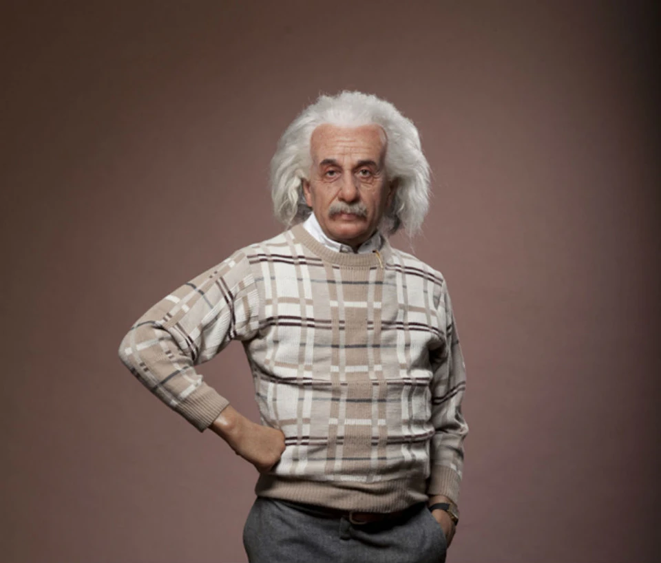 Older man with white hair and mustache, wearing a beige plaid sweater and grey pants, standing against a plain background.