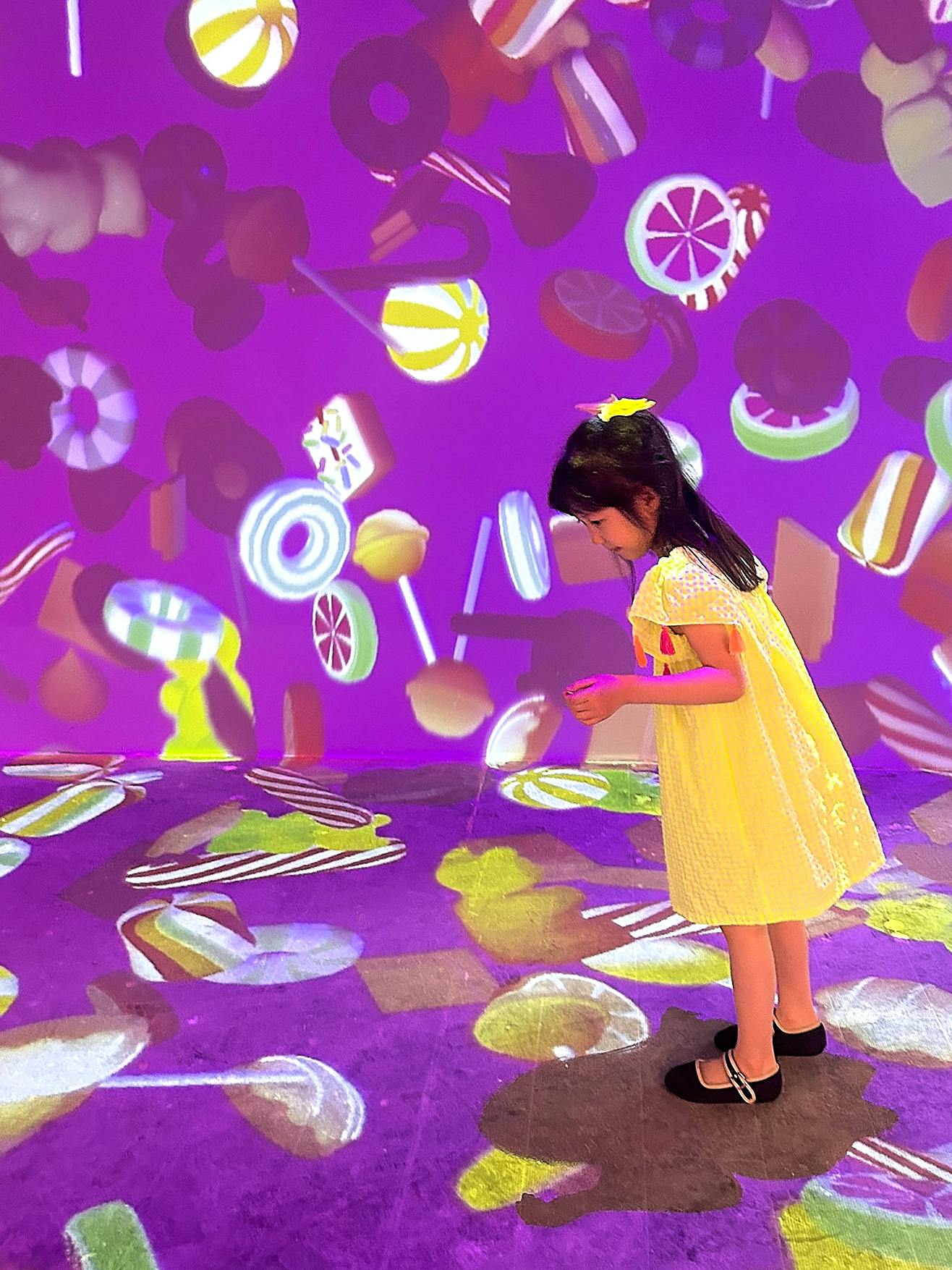 Uma criança com um vestido amarelo está numa sala com projeções coloridas com motivos de doces nas paredes e no chão.