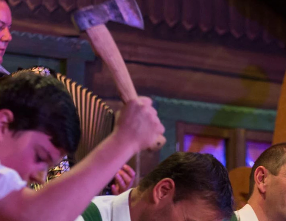 Men in traditional attire perform a log-cutting act on stage, while a musician plays an accordion in the background.