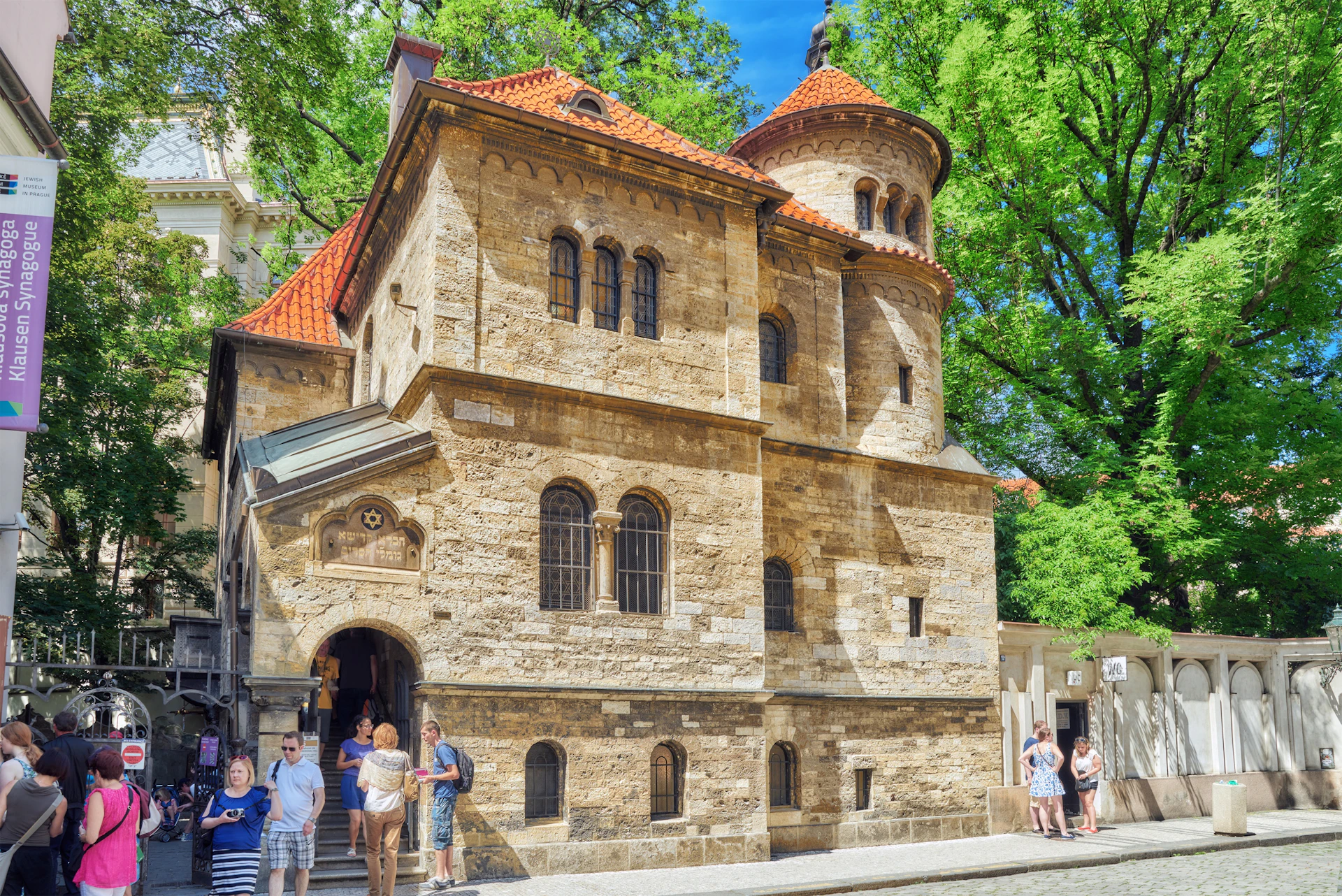 People standing and chatting outside a historic stone building with arched windows and red-tiled roofs, surrounded by trees.