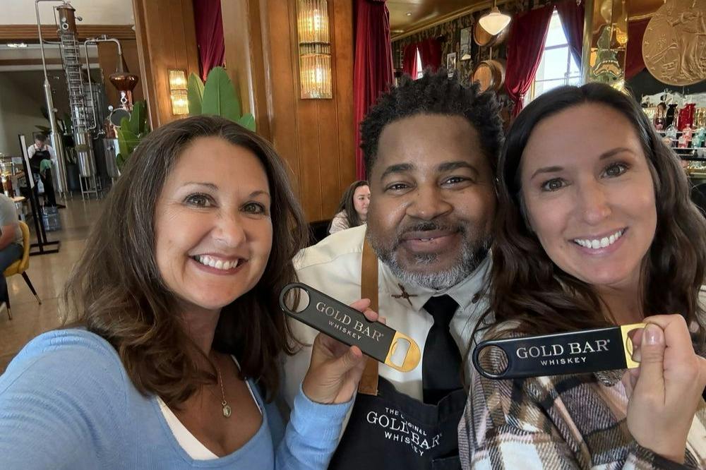 Two women and a man smiling together indoors, holding Gold Bar Whiskey branded items.