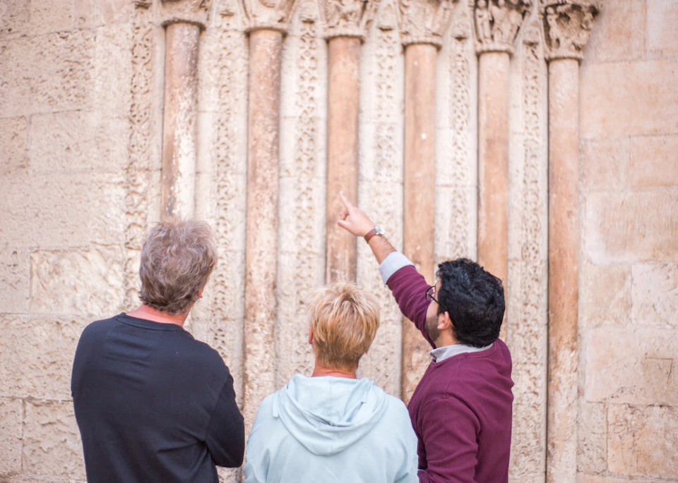 Three people stand in front of an intricately carved stone wall, with one person pointing at the carvings.
