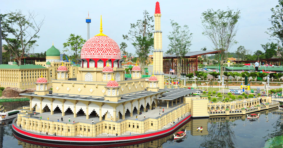 A LEGO model of a mosque with a large red and white dome, minaret, and people figures, set amidst trees and a reflective pond.