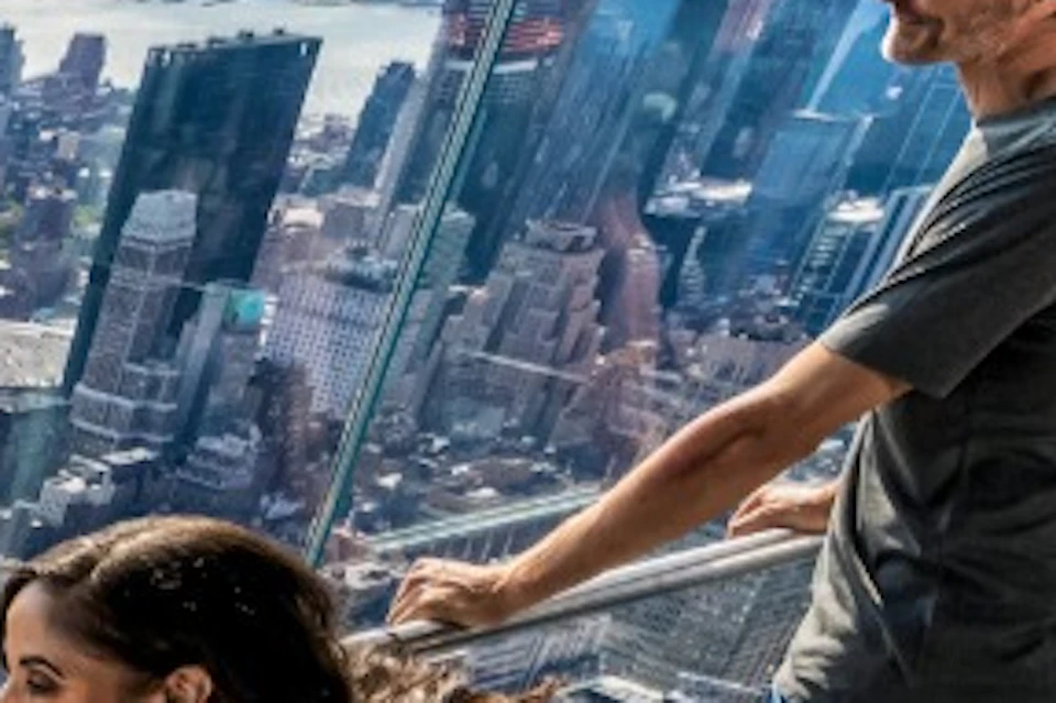 Two people observing a cityscape from a glass observation deck, with a scenic view of buildings and a river in the background.
