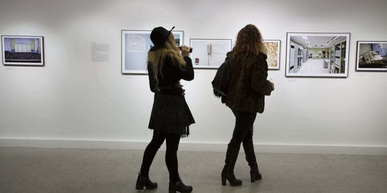 白いギャラリーの壁に額装されたアートワークを見ている2人の女性。