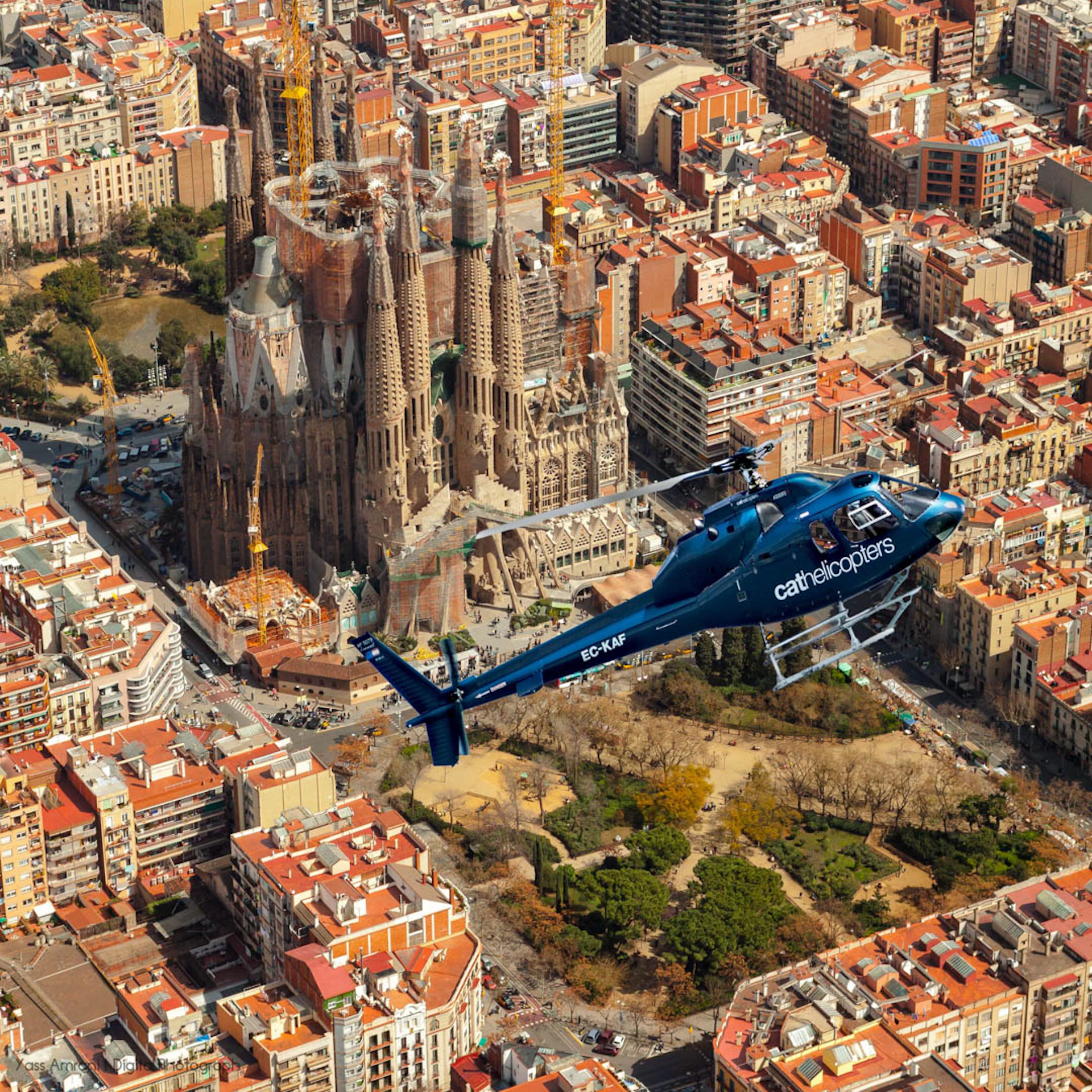 Aerial view of a blue helicopter flying over a dense urban area with the iconic Sagrada Família basilica in the background.