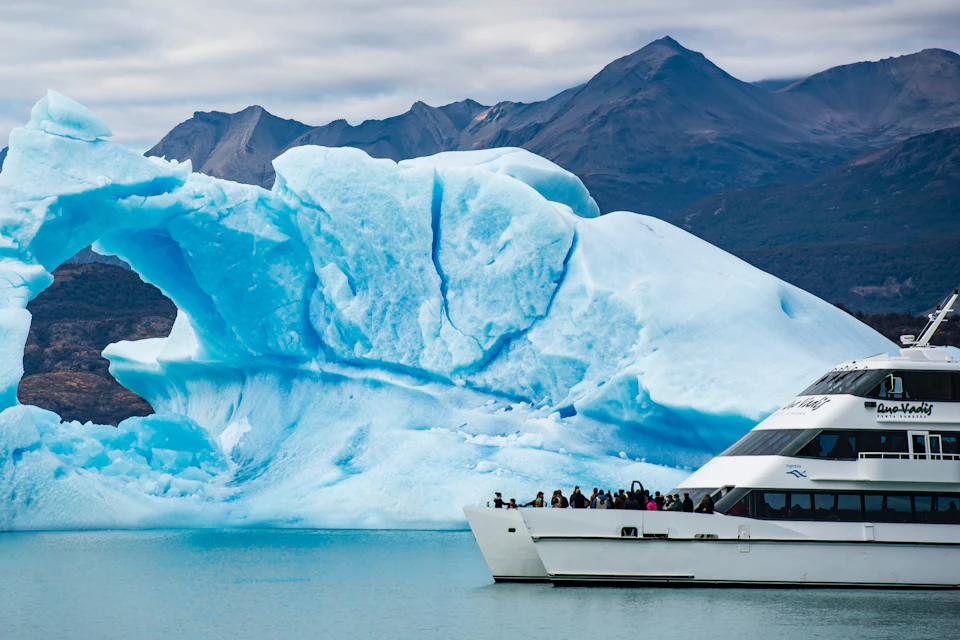 A boat with people onboard approaches a large blue iceberg in a calm body of water, with mountains in the background.