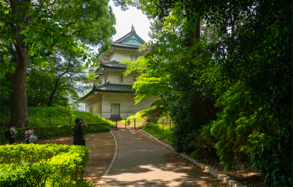 A traditional Japanese structure surrounded by green trees, viewed from a paved pathway with a person taking a photo.
