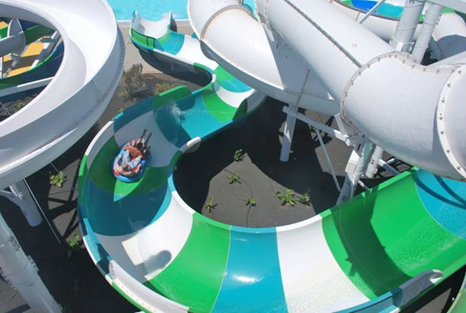 People on a raft slide down a colorful water slide, surrounded by larger white tube slides, with a pool in the background.