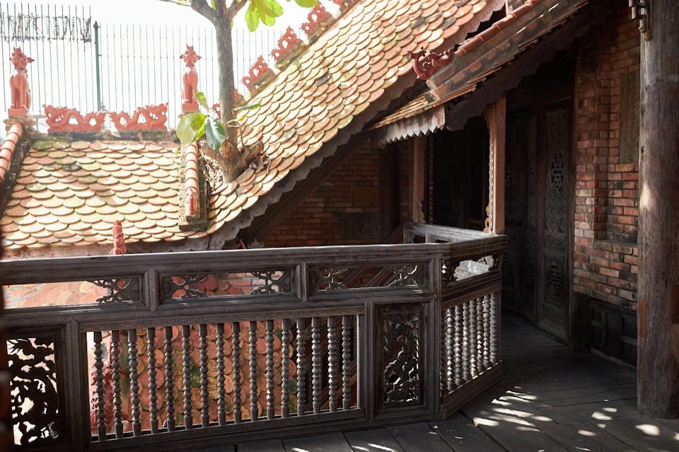 A traditional wooden balcony with intricate carvings overlooks a slanted tiled roof and a tree with green leaves.