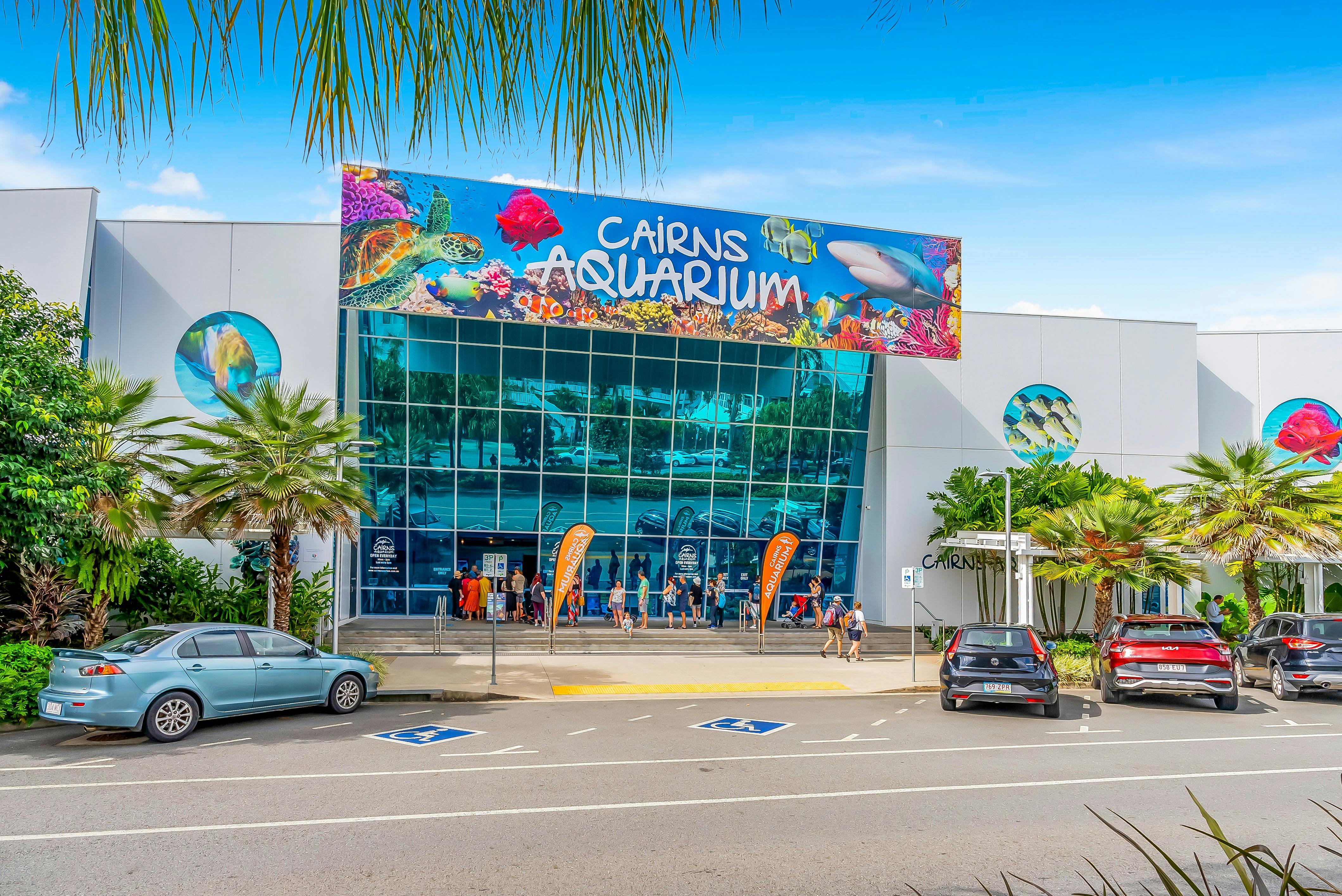 Visitors standing outside Cairns Aquarium entrance with colorful marine life banner, surrounded by tropical plants and parked cars.