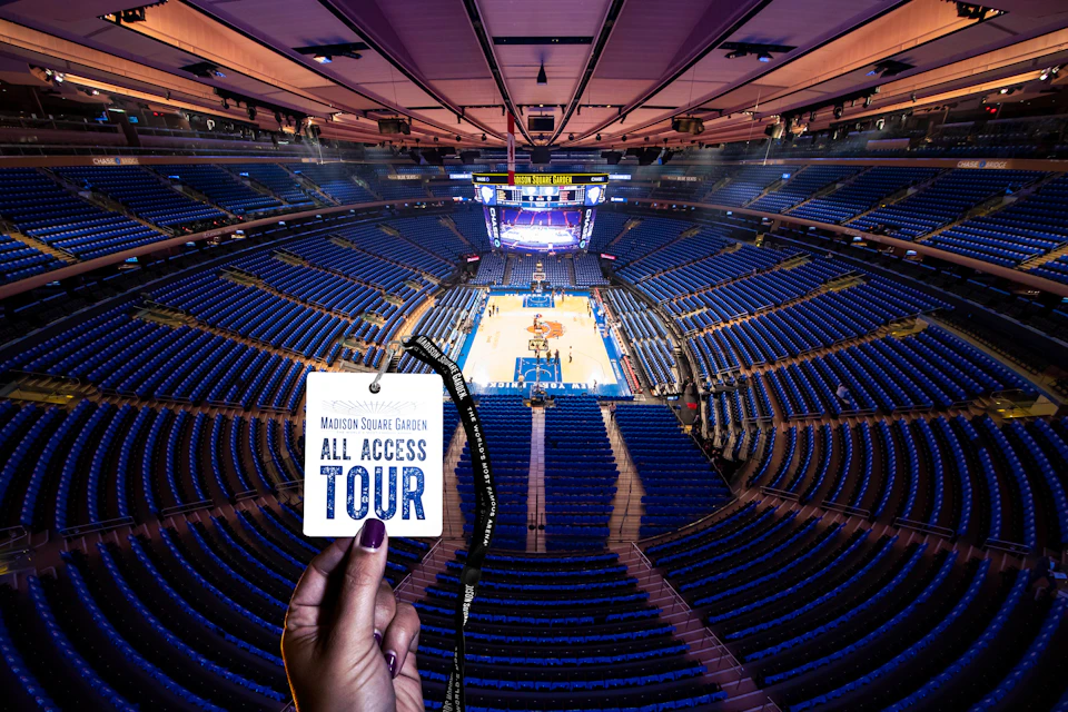 A hand holds an "All Access Tour" pass overlooking an empty Madison Square Garden with a basketball court and blue seats.