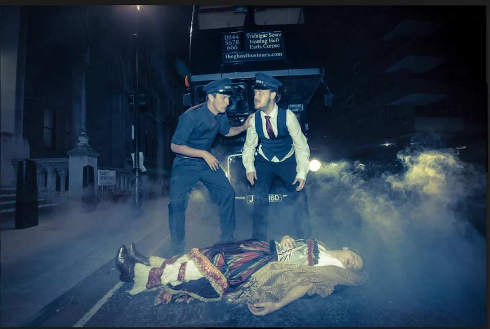 Two men in uniforms stand by a bus, looking at a person in historical clothing lying on the ground in a foggy street at night.