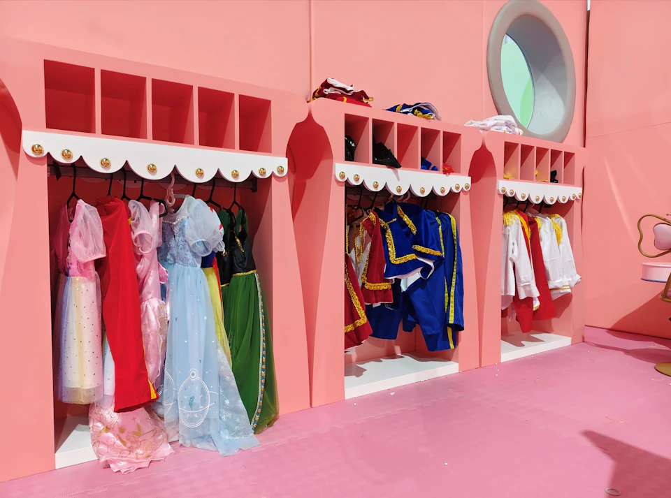 A pink wardrobe with multiple sections holding colorful children's costumes, including princess dresses and regal outfits.