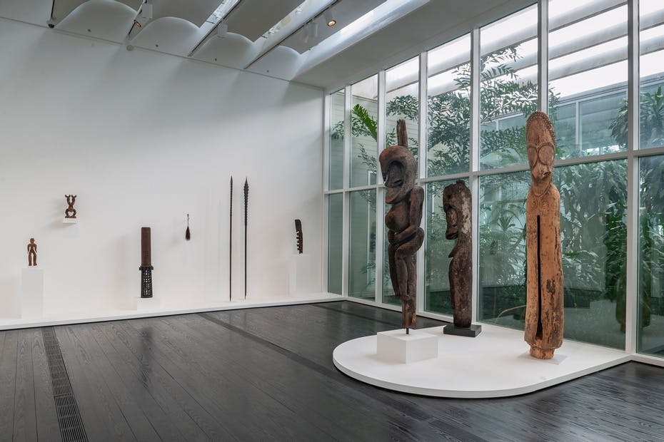 커다란 창문이 있는 갤러리로, 흰색 받침대와 벽에 나무 조각과 높고 날씬한 유물을 전시합니다.