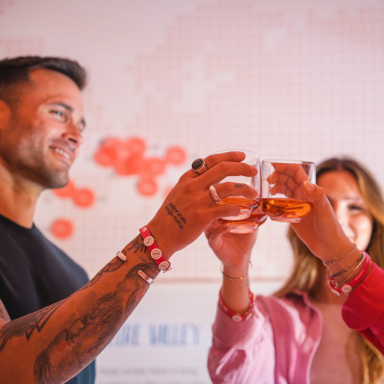 Three people raising glasses in a toast, wearing colorful bracelets, with a blurred wall map in the background.