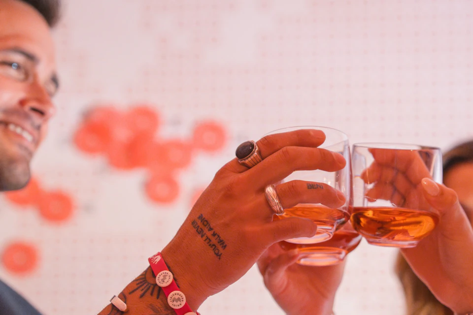 Three people raising glasses in a toast, wearing colorful bracelets, with a blurred wall map in the background.