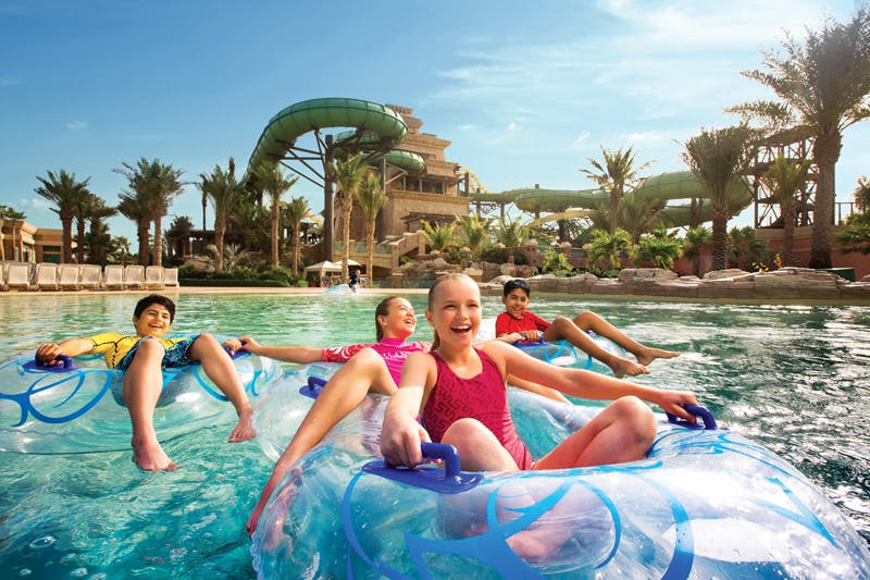 Children floating in clear inner tubes in a water park, smiling and enjoying the ride. Water slides and lush greenery in the background.