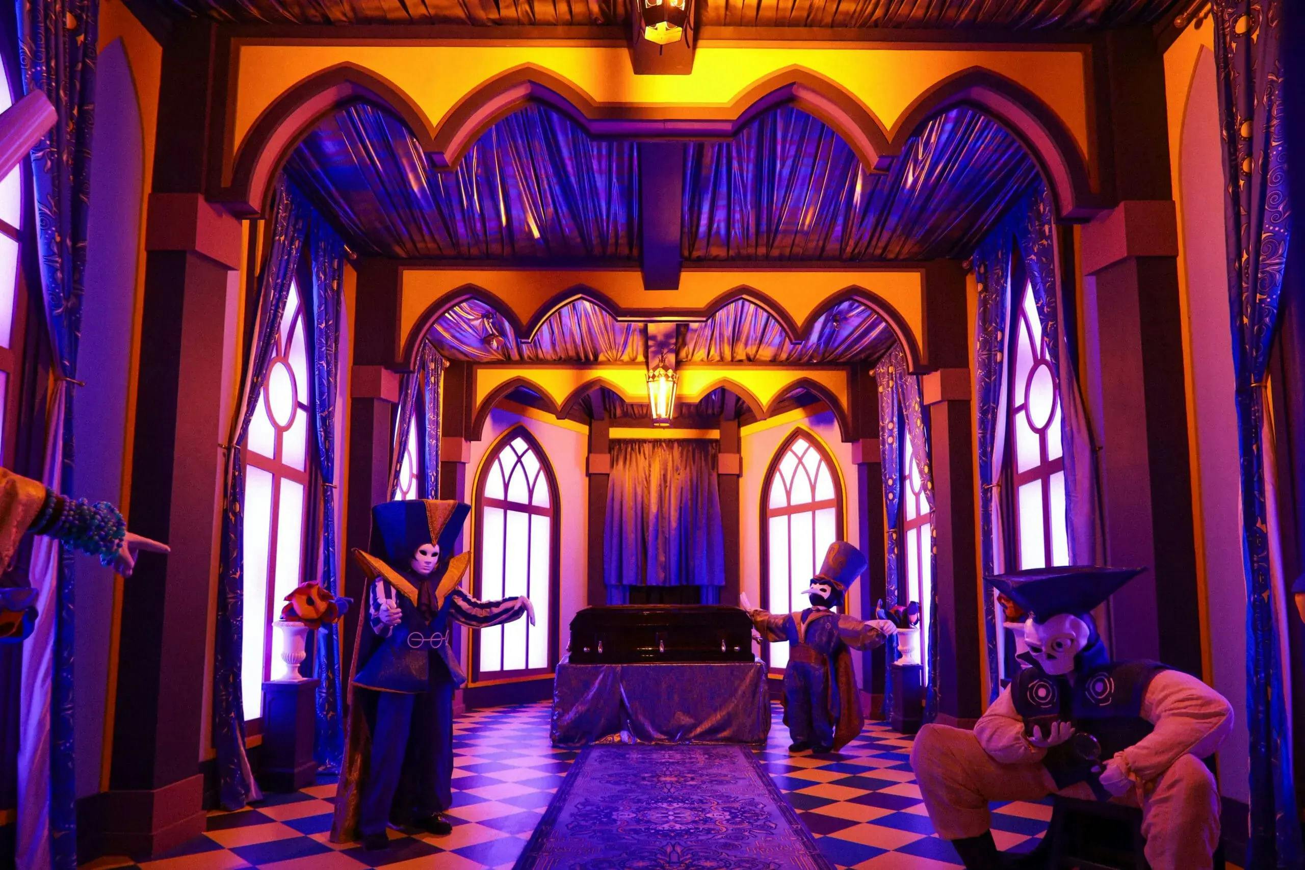 A room with arched windows, blue lighting, and three people in colorful costumes and masks, arranged around a covered piano.