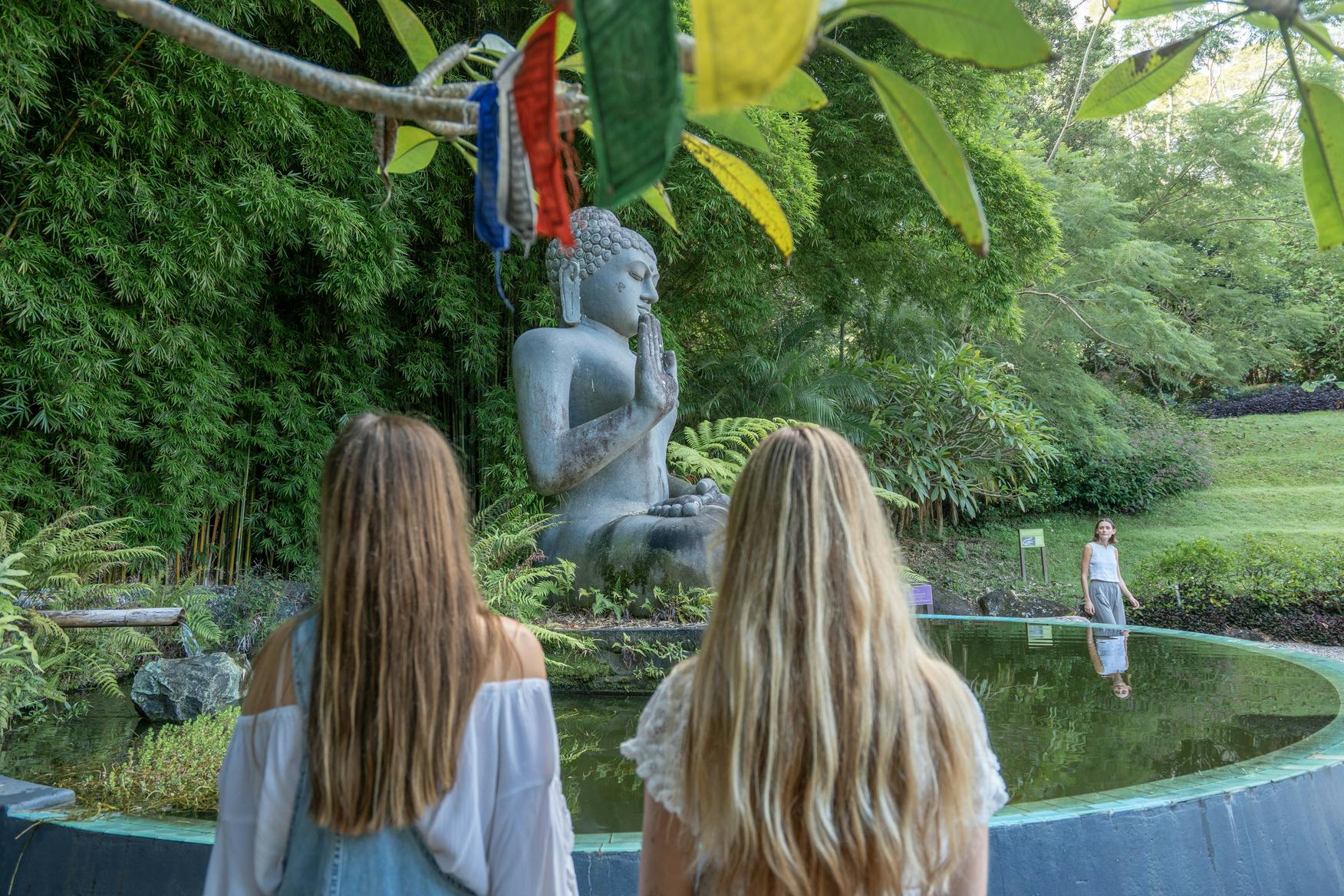 緑と祈りの旗に囲まれた池の近くにある石造りの仏像に面した長い髪の女性2人が立っています。