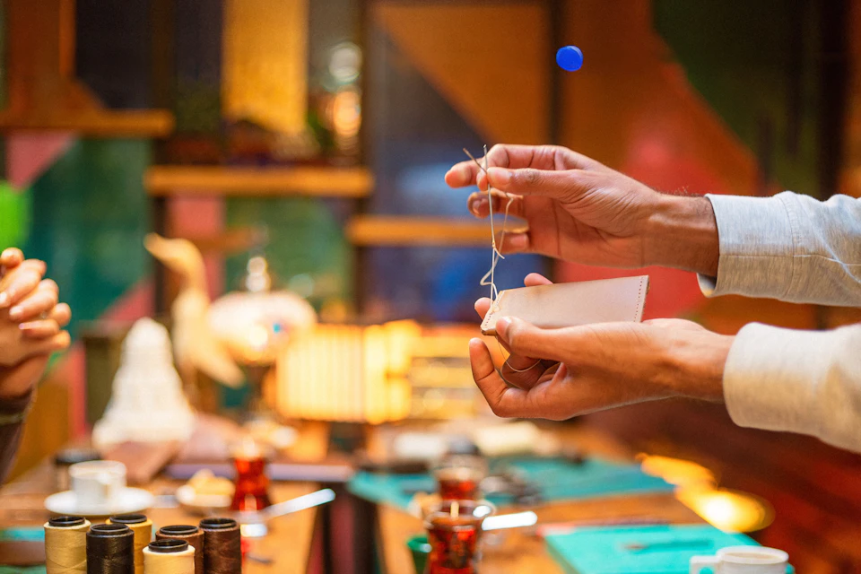 Close-up of hands stitching leather with a needle and thread in a colorful workshop setting, tools visible on the table.