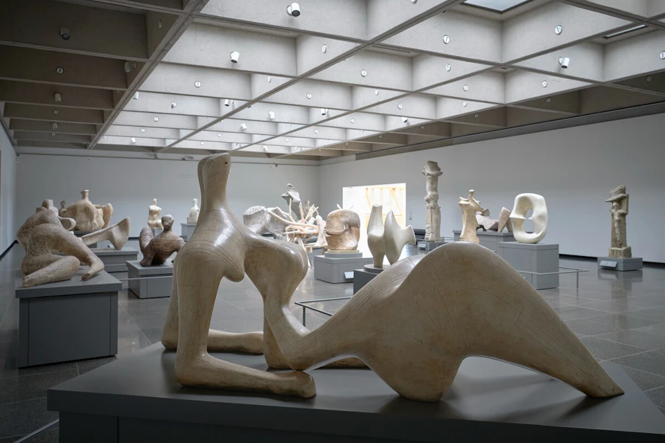 A modern art gallery with various abstract sculptures displayed on pedestals under a grid-patterned ceiling.